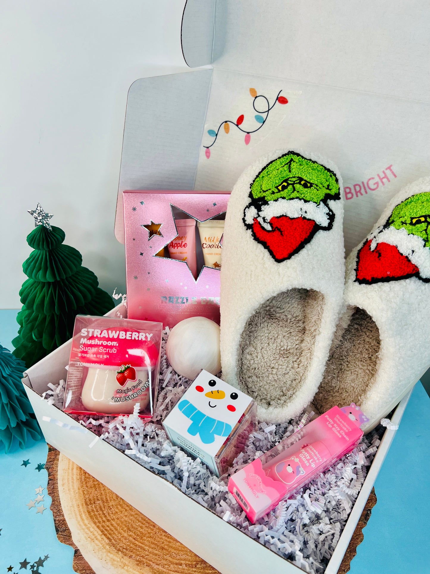A holiday-themed gift set featuring a Grinch-themed slipper, strawberry shortcake lip gloss, a snowball surprise, hand cream gift set, and gummy candies, all arranged in a gift box with holiday decorations.