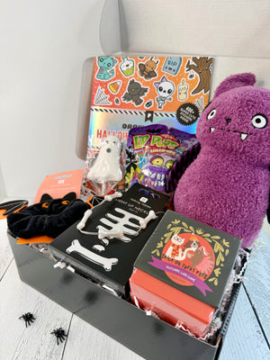 A Halloween-themed gift box filled with various spooky treats and accessories, including a purple plush toy, a bag of purple candy, a box of 'Spooktacular' popcorn, a black pair of clawed gloves, and a sticker sheet.