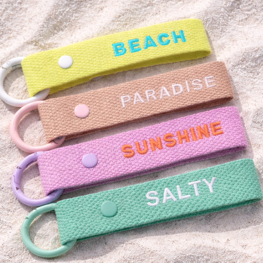 Four colorful keychains with embroidered text on a sandy background