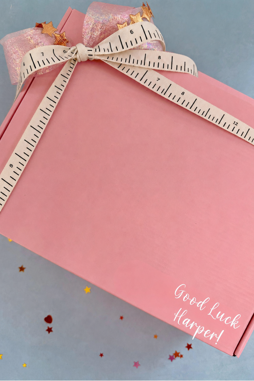 A pink gift box tied with a cream measuring tape ribbon and bow, decorated with small gold stars, on a light blue background with scattered star confetti. The box has the words 'Good Luck Harper!' printed in white script.