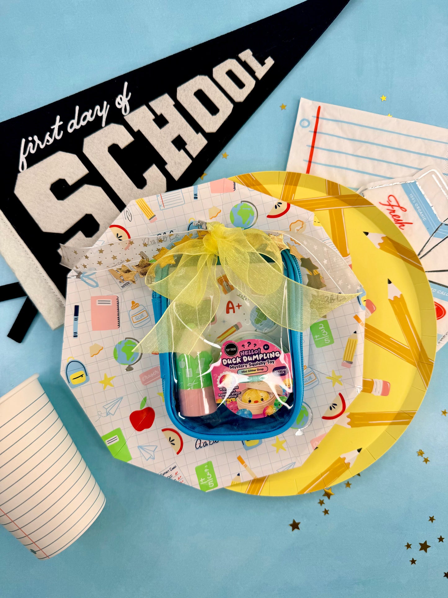 A first day of school gift basket arrangement on a light blue background, featuring a blue pencil case with a yellow ribbon, a pink and white 'Pencil Push Pin' magnet, school-themed paper plates, a black pennant reading 'first day of SCHOOL', and lined paper props.