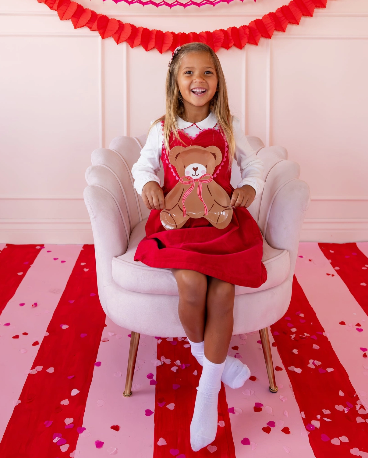 Young girl in a red dress holding a teddy bear, sitting on a pink chair with a heart-themed floor and backdrop.