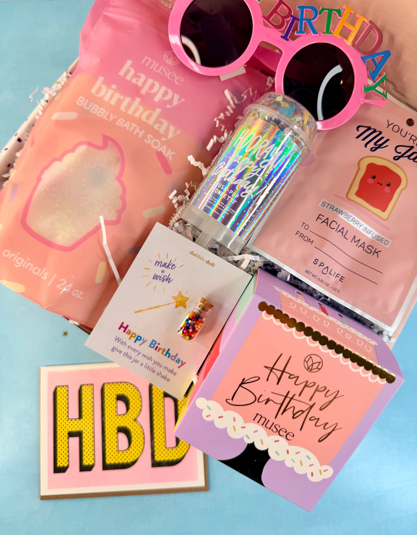 A birthday gift set arranged on a light blue background, featuring a pink bag of musce bubble bath soap, pink novelty sunglasses, a holographic tumbler bottle, a pink facial mask card, a small colorful bead vial on a card, and multiple pink and lavender birthday cards and stickers.