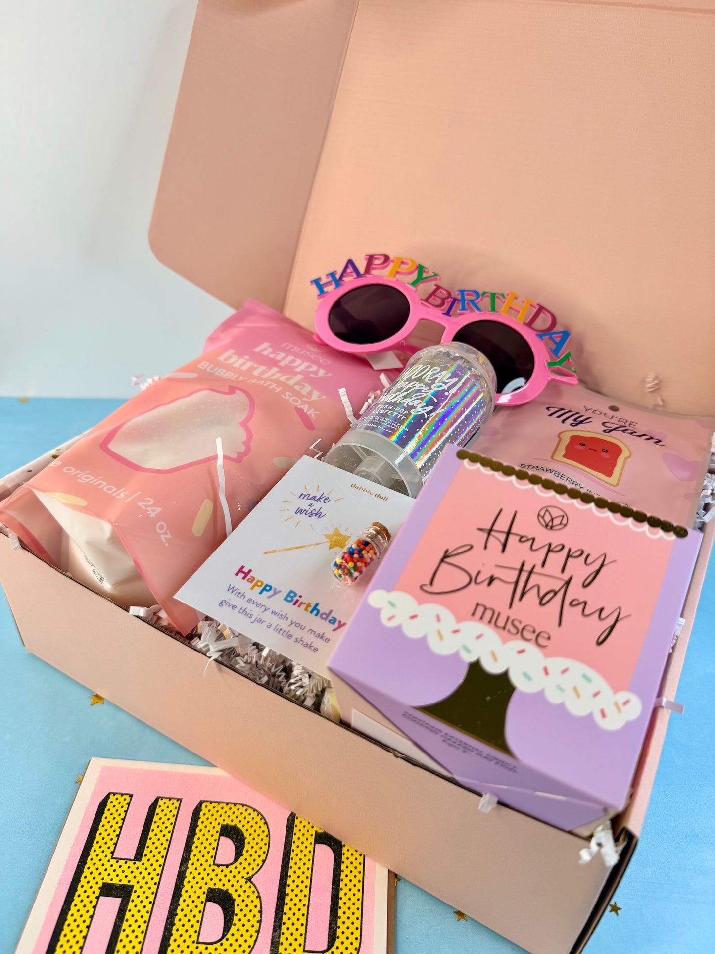 A pink gift box filled with birthday-themed items, including pink novelty sunglasses, a pink bag, a holographic bottle, a purple card, and a pink card reading 'Happy Birthday musee', with a matching 'HBD' card in front.
