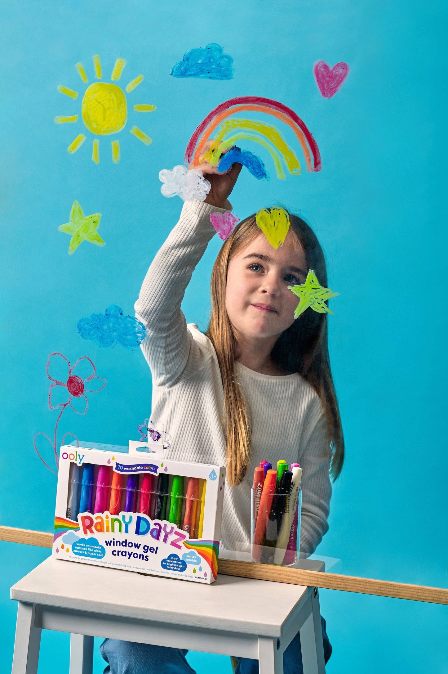 Child with colorful window gel crayons and a box of Crayola paint, against a blue background with rainbow and cloud decorations.