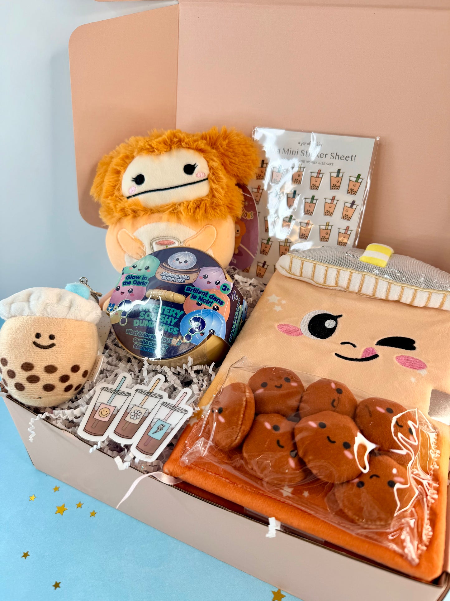 A curated snack box gift set filled with cute plush toys and stationery. The set includes an orange lion plush, a brown bubble tea plush, a round tin of bubble tea charms, a mini sticker sheet, and a plush cookie bag filled with brown plush cookies, all arranged in a cardboard box.
