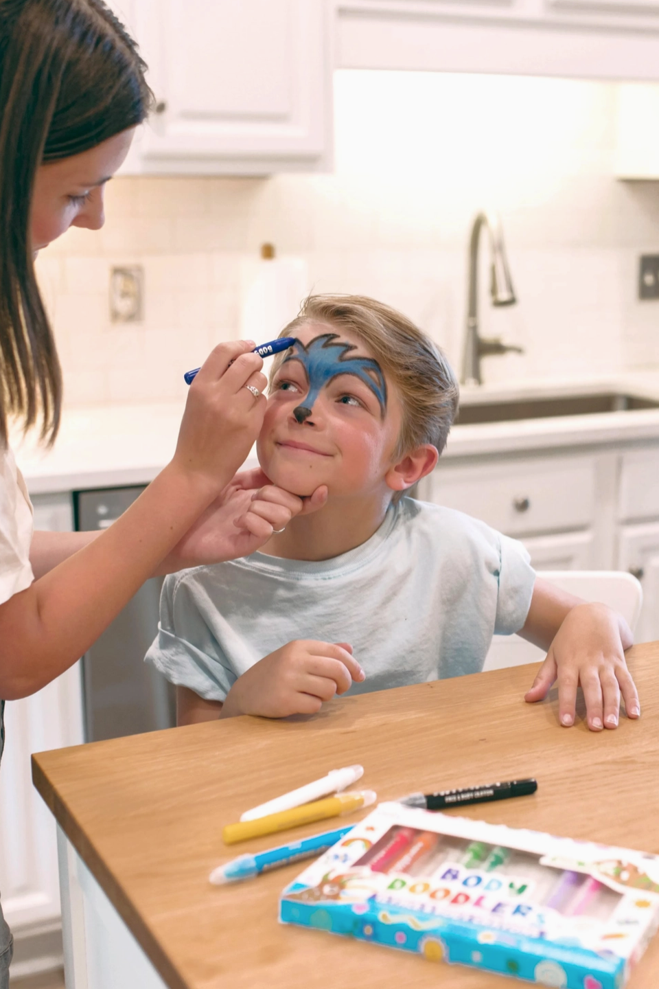 Person applying face paint to a child's face in a kitchen setting