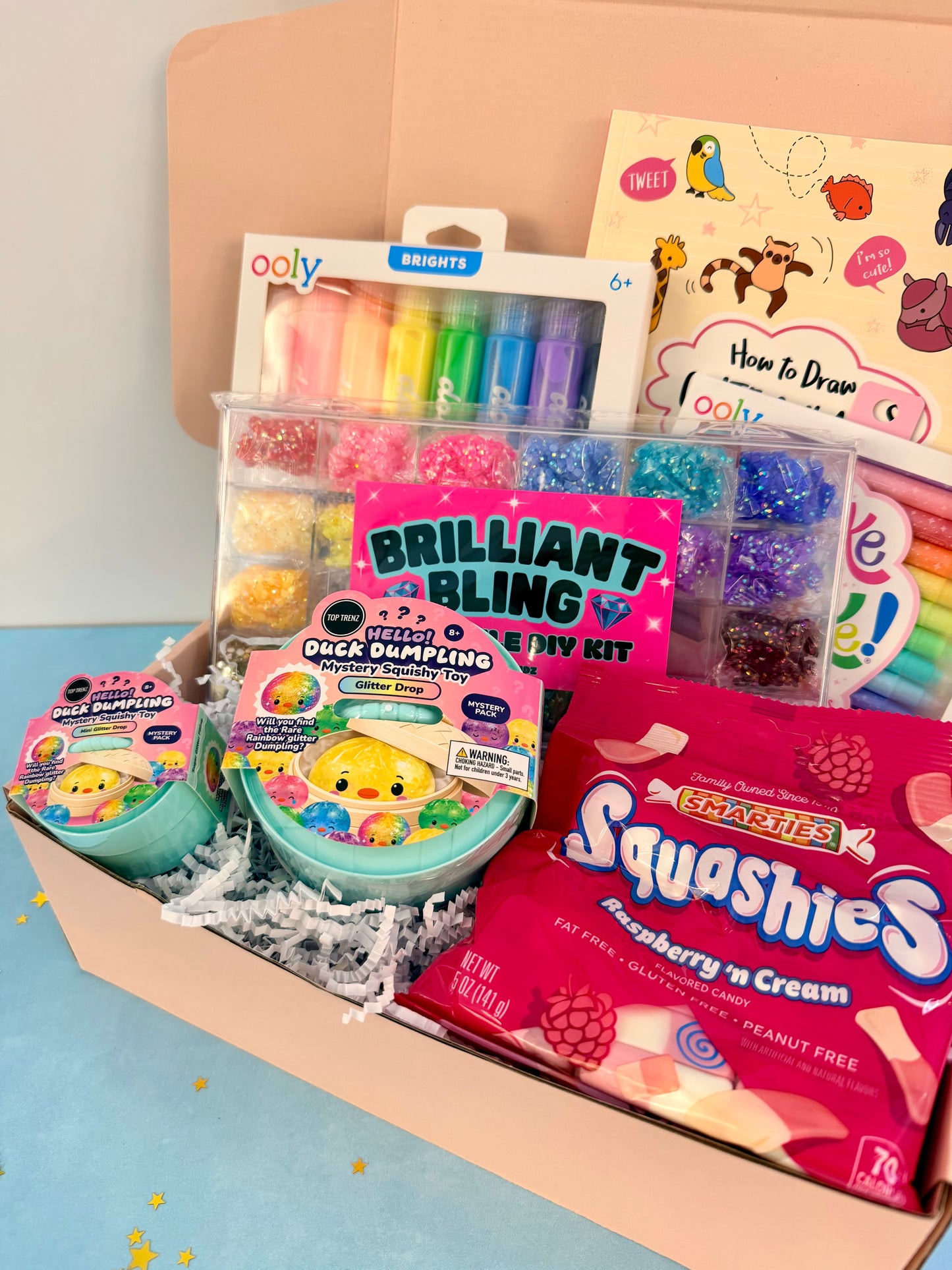 A curated gift box filled with colorful craft and play items, including a rainbow marker set, a glitter tray, a pink 'Brilliant Bling' daily kit card, a round toy container, a bag of Mash & Pies Squashies candy, and a drawing guide booklet.