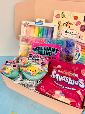 A curated gift box filled with colorful craft and play items, including a rainbow marker set, a glitter tray, a pink 'Brilliant Bling' daily kit card, a round toy container, a bag of Mash & Pies Squashies candy, and a drawing guide booklet.