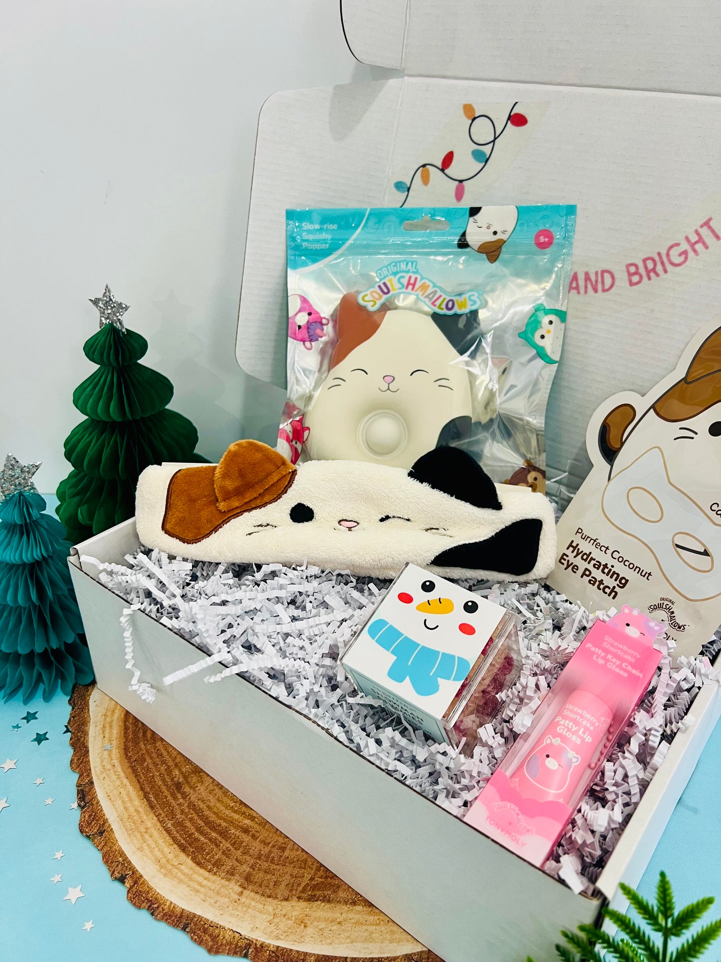 A gift box with a Squishmallow Cloud Pop, Squishmallows X TONYMOLY sheet mask, Squishmallows X TONYMOLY keychain lip gloss, and Sugarfina Snowman Gummies, decorated with a Christmas tree and ornaments.