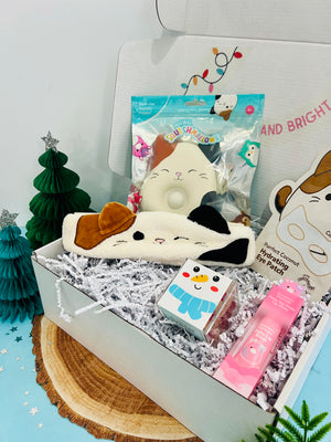 A gift box with a Squishmallow Cloud Pop, Squishmallows X TONYMOLY sheet mask, Squishmallows X TONYMOLY keychain lip gloss, and Sugarfina Snowman Gummies, decorated with a Christmas tree and ornaments.