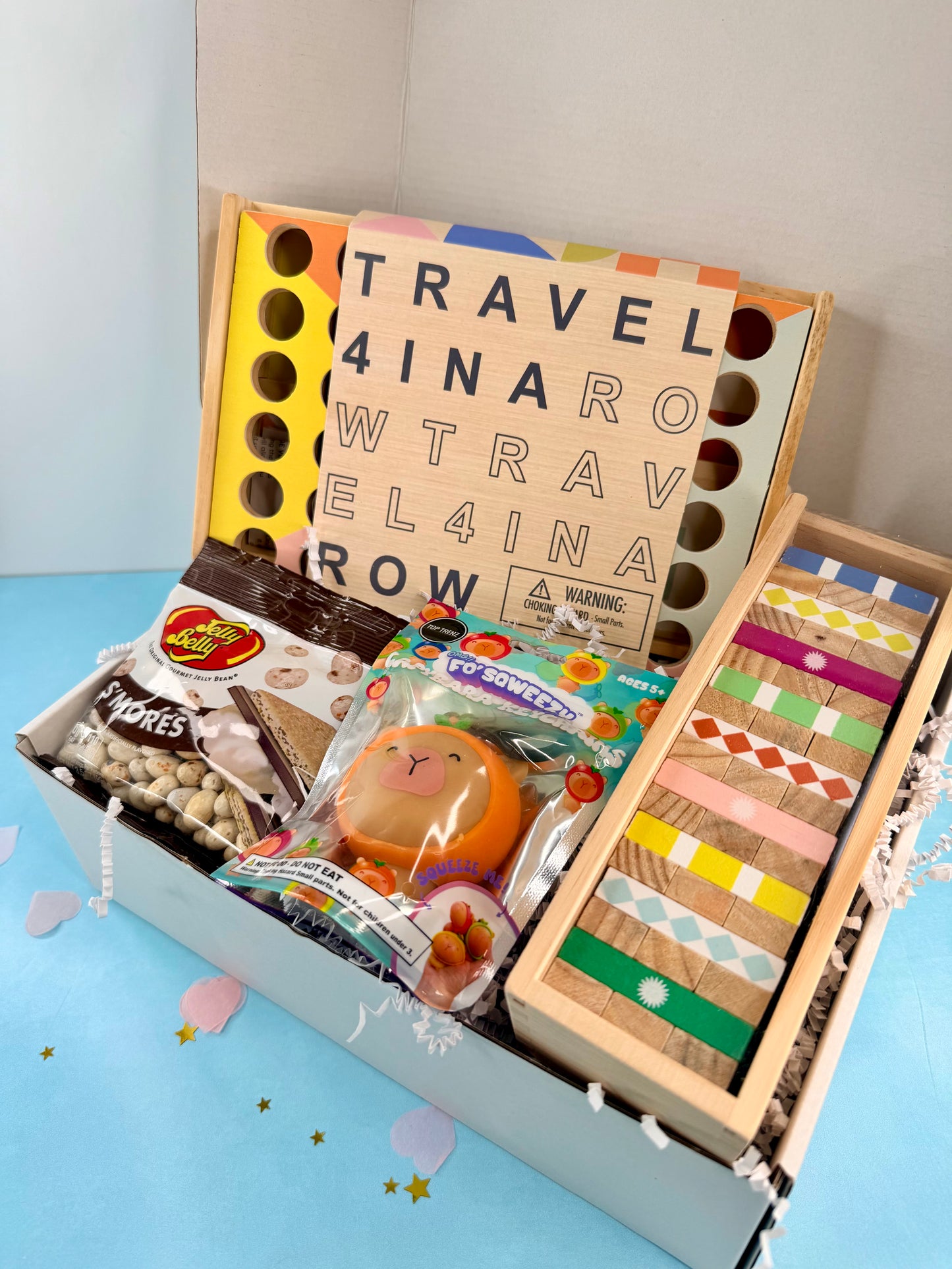 Travel-themed gift box with wooden game, snacks, and toys on a light blue surface.