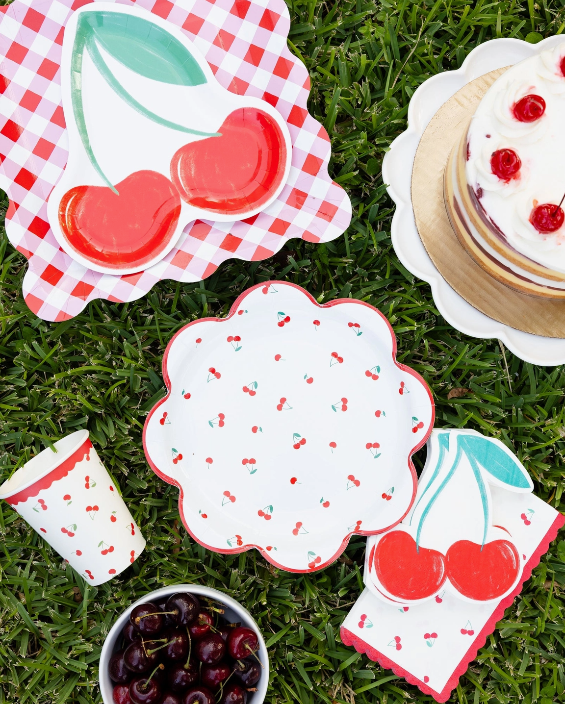 Set of picnic-themed plates, cups, and napkins with cherry design on grass.