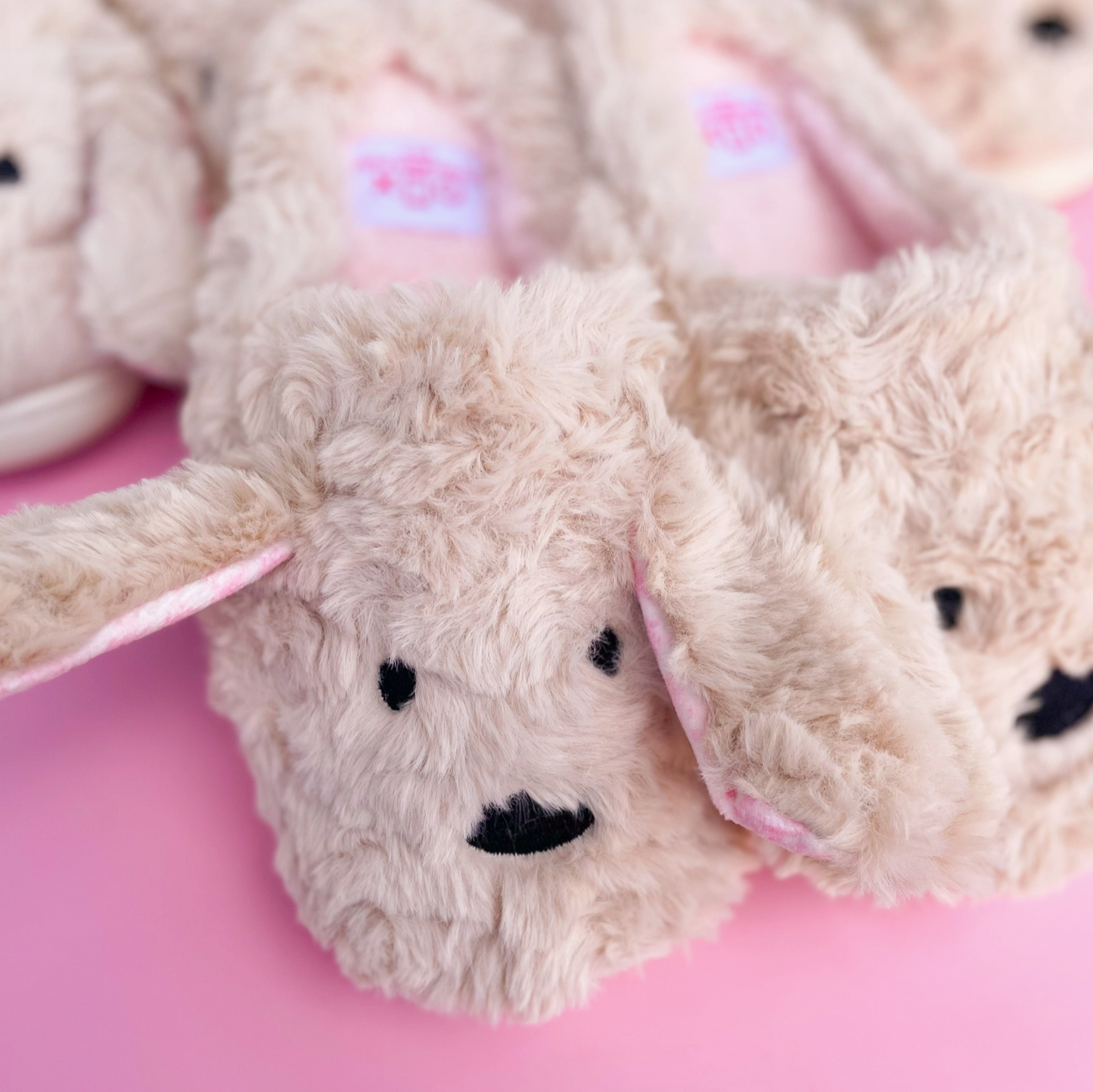 A pair of beige plush dog slippers with floppy ears and a smiling face design on a pink background.