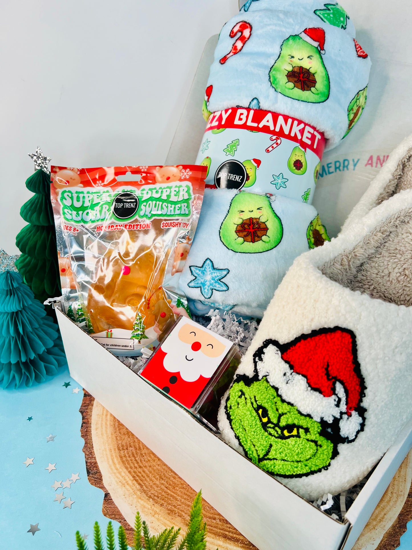 A gift box with a 'Super Duper Sugar Squisher - Reindeer' and an 'Avocado Xmas Fuzzy Throw Blanket' with The Grinch design on it, along with 'Sugarfina Santa Holiday Trees' and a green Christmas tree ornament.