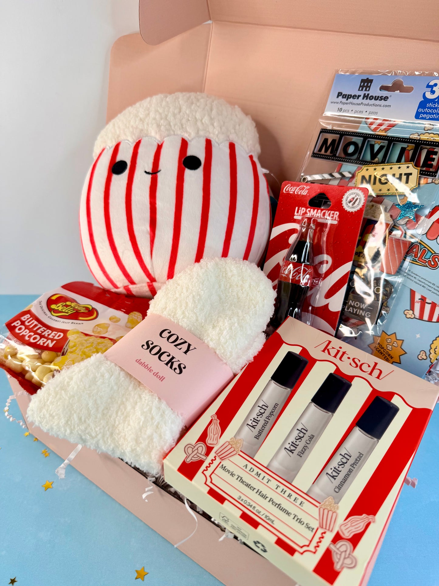A curated gift box filled with themed items, including a plush character in red and white stripes, a pink sock, a KitSch perfume set, a bag of Pop-Tart, and a bag of Kicker's Butter Popcorn, all arranged in a pink box.