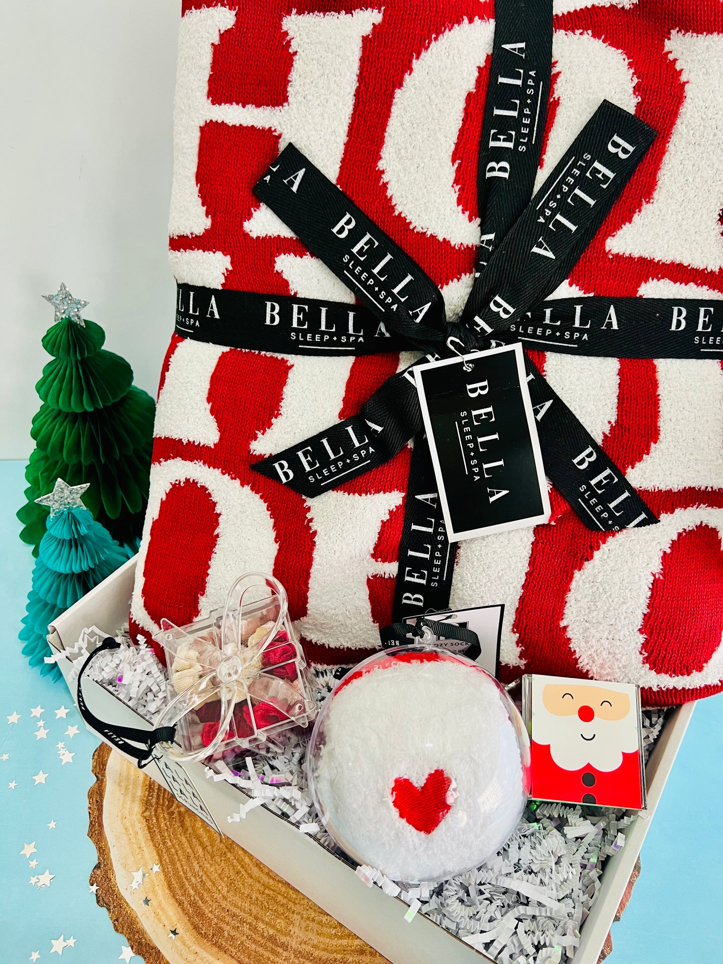 A holiday gift box containing a cozy red and white checkered throw, a pair of cozy red socks with a white heart, and a velvet scrunchie present. The gift is decorated with a 'La Bella' brand tag and a Santa Claus card, presented against a backdrop of a Christmas tree and holiday decorations.