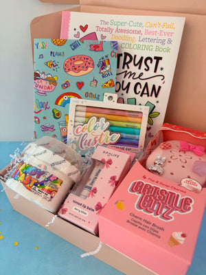 A curated gift box filled with stationery and beauty items, including a colorful coloring book, a pink hair brush box labeled 'Grasshopper Blitz,' lip balm, markers, and small packaged accessories, arranged in a light wood box on a pastel blue surface.