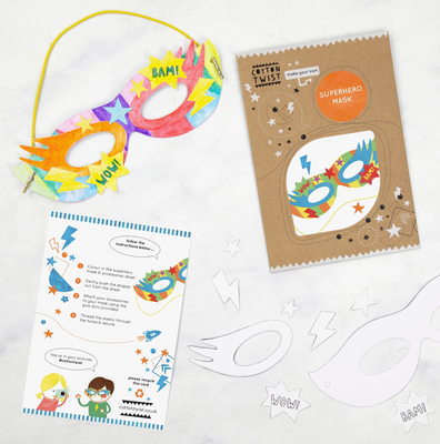 A colorful superhero mask with yellow straps and a cardboard base, alongside a kraft envelope with an instruction card and glue dots.