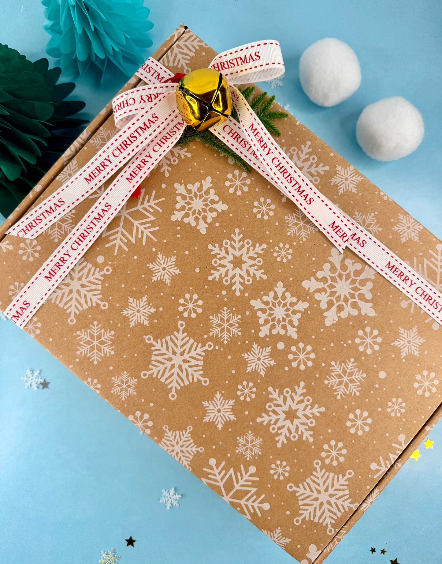 Gift box wrapped in brown paper with white snowflake pattern, tied with a red ribbon and gold jingle bell on a blue background.