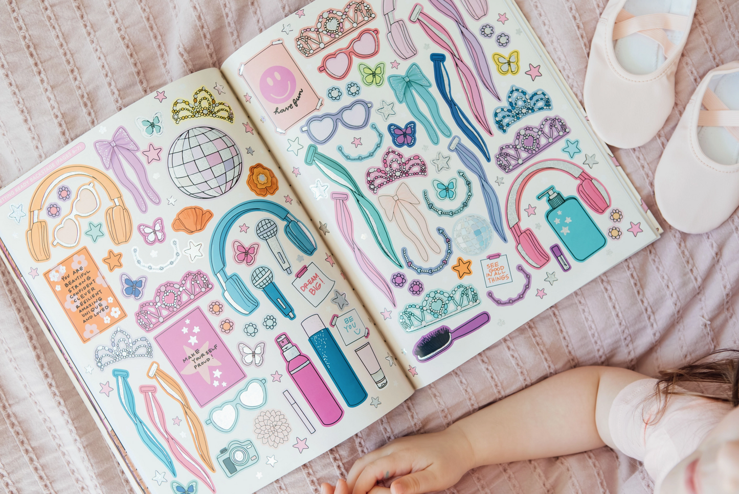 Colorful sticker book open on a pink blanket with a child's hand reaching towards it.