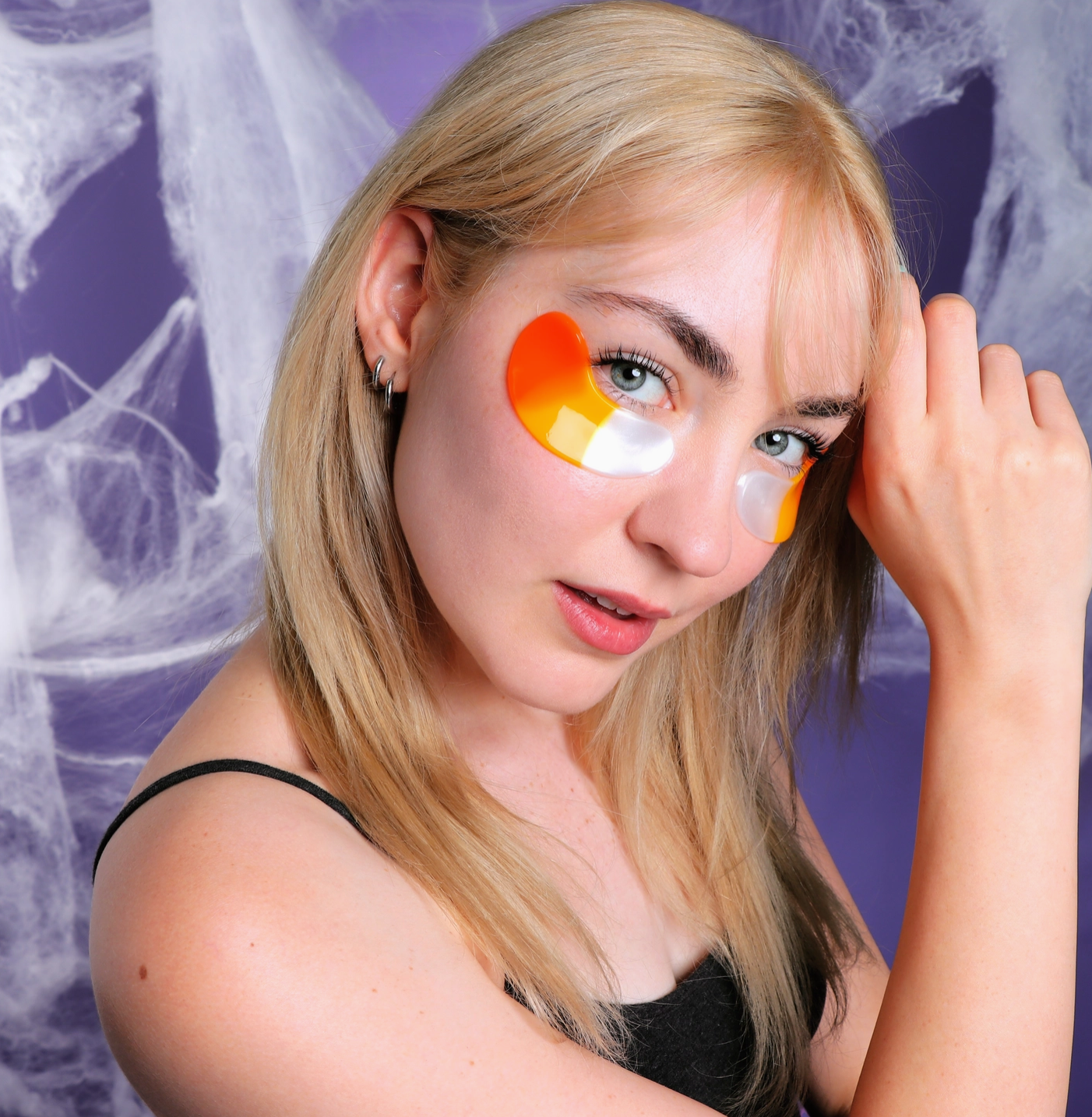A blonde woman wearing orange and white crescent-shaped eye patches, posing against a purple background with white spiderwebs.