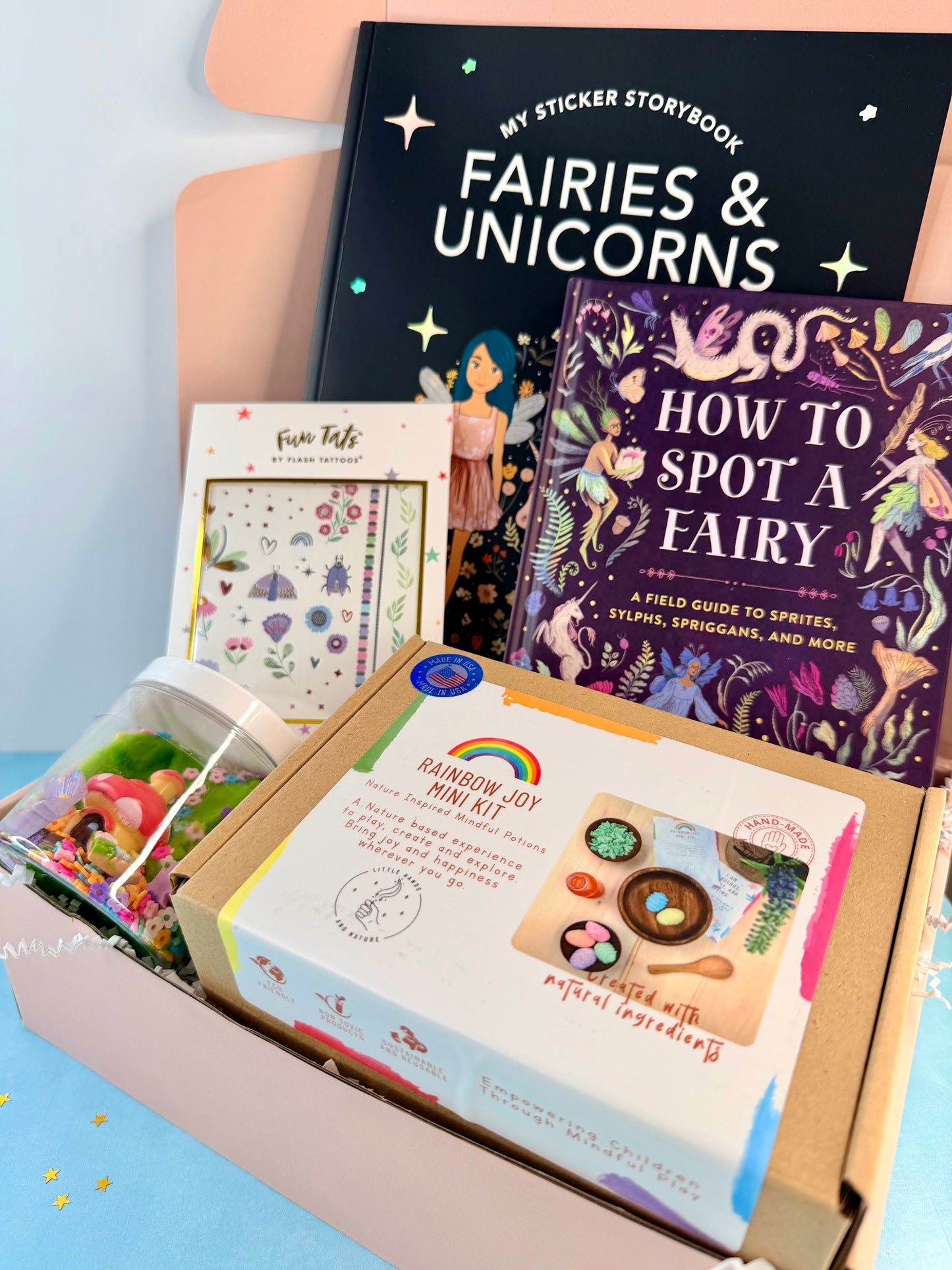 A curated fairy-themed gift box or kit display featuring a black 'Fairies & Unicorns' sticker storybook, a purple 'How to Spot a Fairy' guide, a colorful 'Rainbow Joy Art Kit' box, a 'Fun Facts' card, and a bag of craft supplies inside a pastel pink box.