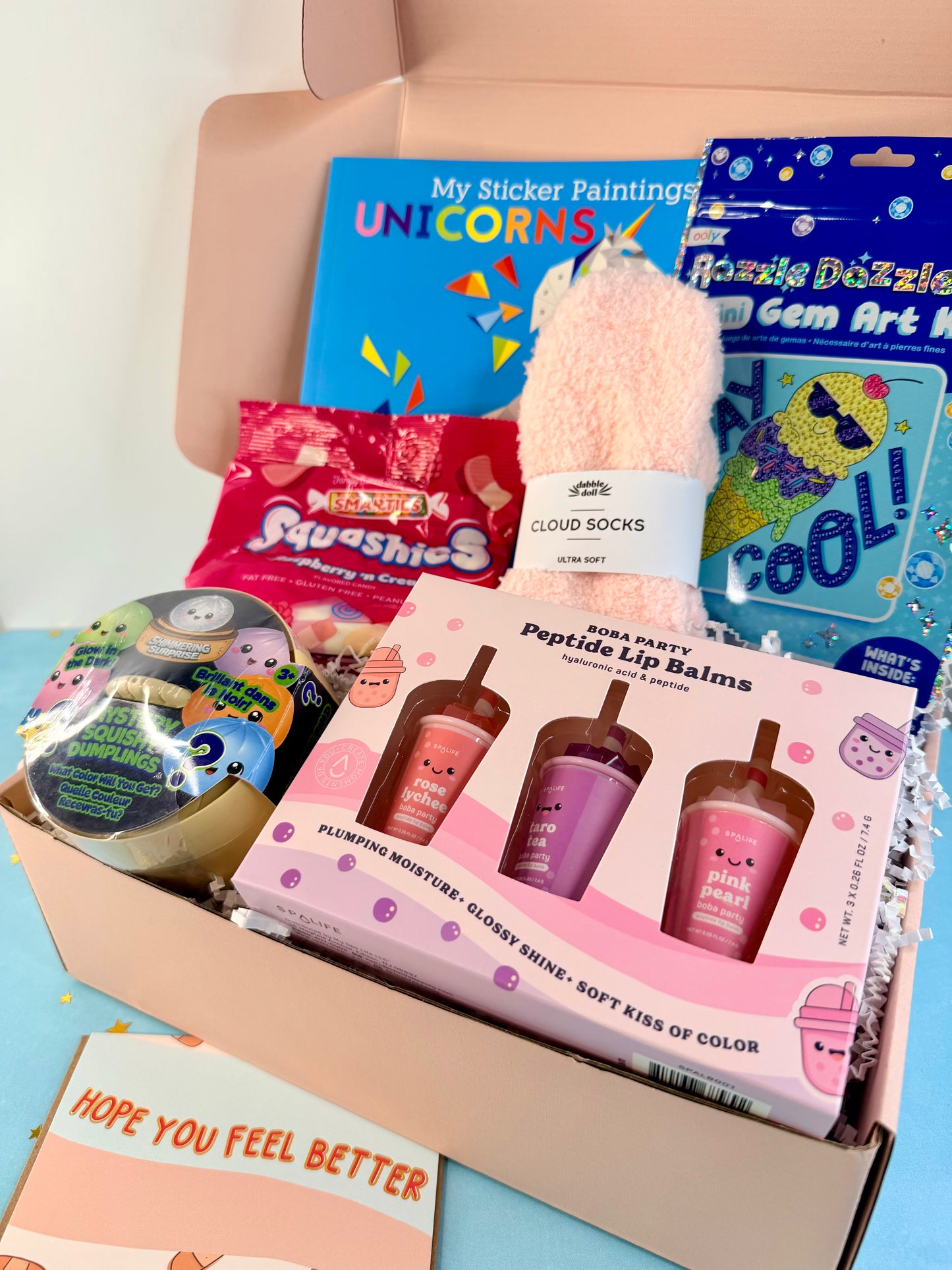 A curated gift box filled with various colorful items, including a pink fuzzy sock, a box of Peppy Peppy Peppermint Lip Balm, a sticker painting book, a gem art kit, and a Squishies bag, arranged in a light brown cardboard box with shredded paper filler.