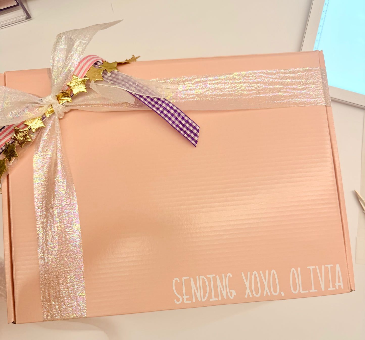A peach-colored corrugated gift box tied with a white iridescent ribbon, gold tinsel, and a purple gingham accent ribbon. The box has 'SENDING XOXO, OLIVIA' printed in white text in the bottom right corner.