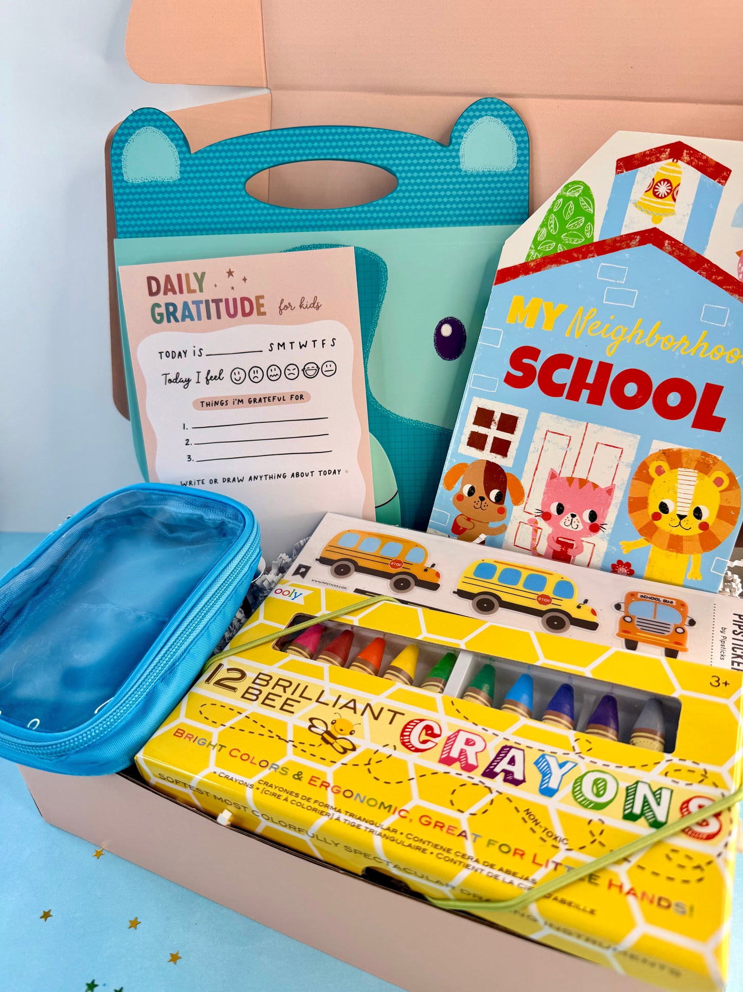 A curated children's gift box or set arranged in a cardboard tray, featuring a teal bear-shaped pouch, a blue zip pouch, a yellow crayon box labeled '12 BRILLIANT BEE CRAYONS,' a colorful children's book titled 'My Neighborhood School,' and a white daily gratitude card.