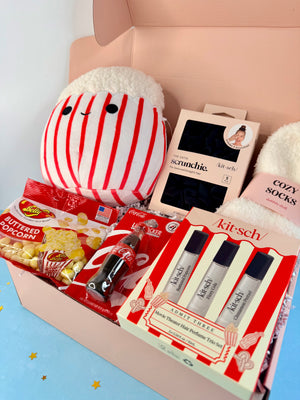 A curated gift box filled with various items including a red and white striped plush toy, a boxed hair treatment set, a pair of cozy socks, and bags of popcorn and candy, all arranged inside a light pink box.