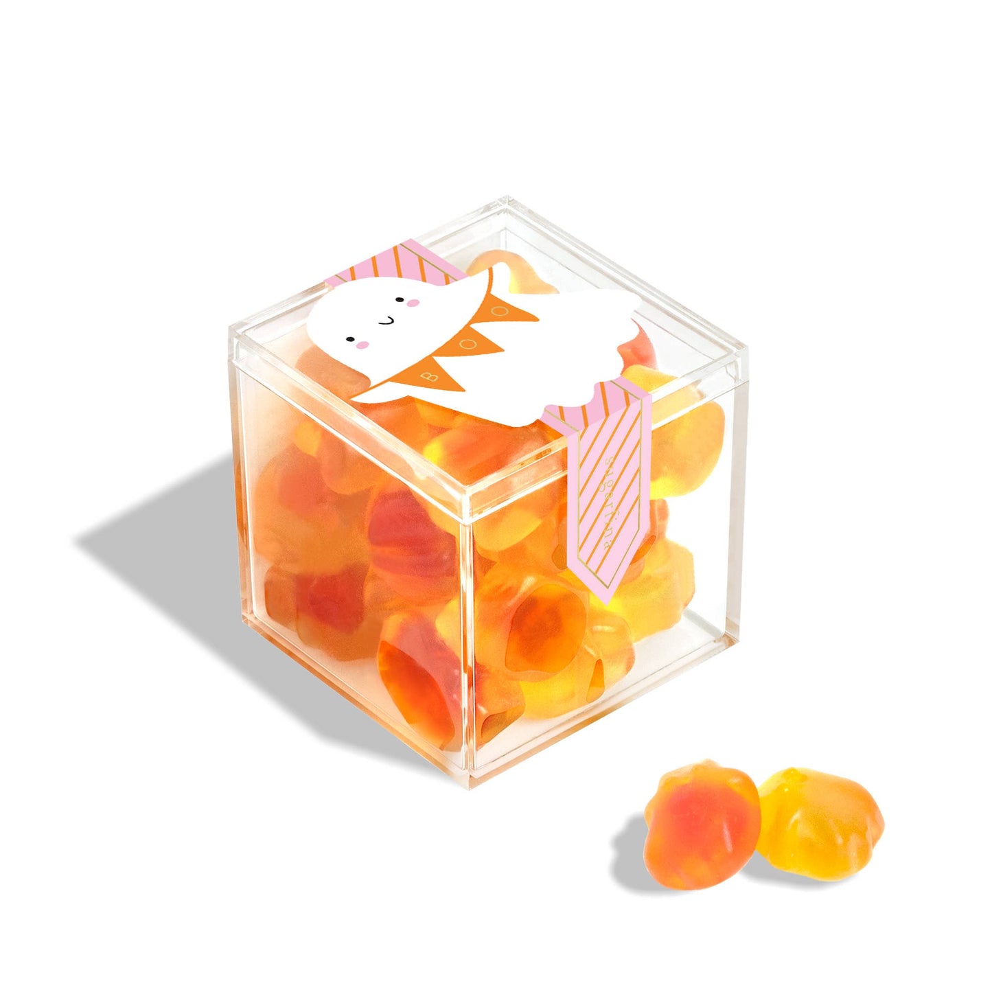 A clear acrylic cube container filled with orange and yellow gummy candies, decorated with a cute white cartoon character wearing a pink striped tie. Two loose candies sit in front on a clean white background.