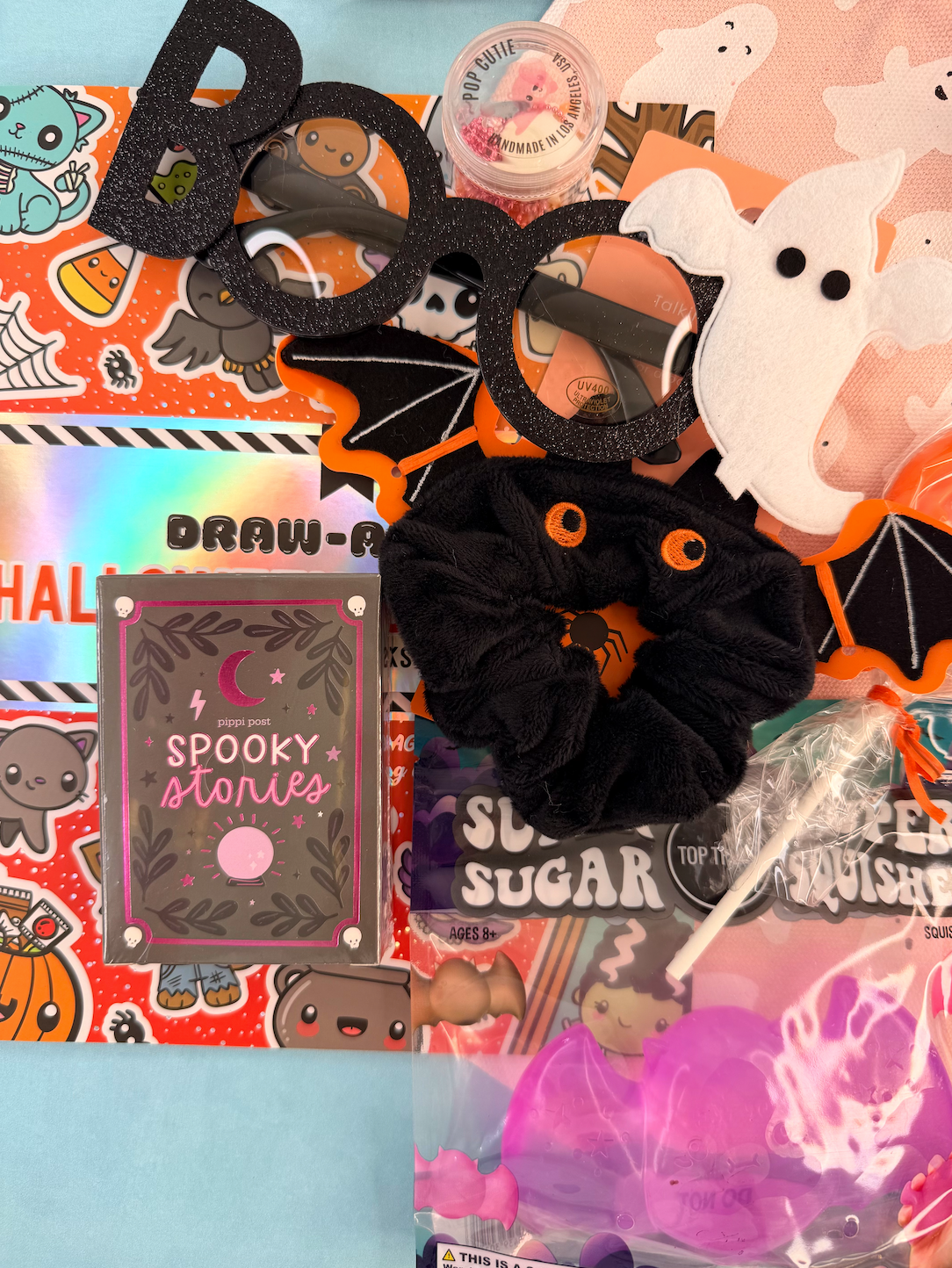 A flat-lay collection of Halloween-themed stationery and accessories, including black glittery 'BOO' letter glasses, a black plush scrunchie with orange eyes, a 'Spooky Stories' notebook, a 'Draw-A-Princess' sticker sheet, and a pink jellyfish lollipop in a clear bag.