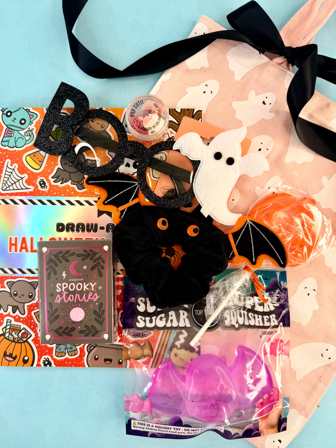 A Halloween-themed squishy and accessory bundle laid out on a light blue background, including a pink ghost-print drawstring bag, black glittery BOOC letter headbands, a black bat-shaped scrunchie, a white ghost plush, orange squishies, a purple bat-shaped item in a bag, stickers, and a 'Spooky Stories' card.