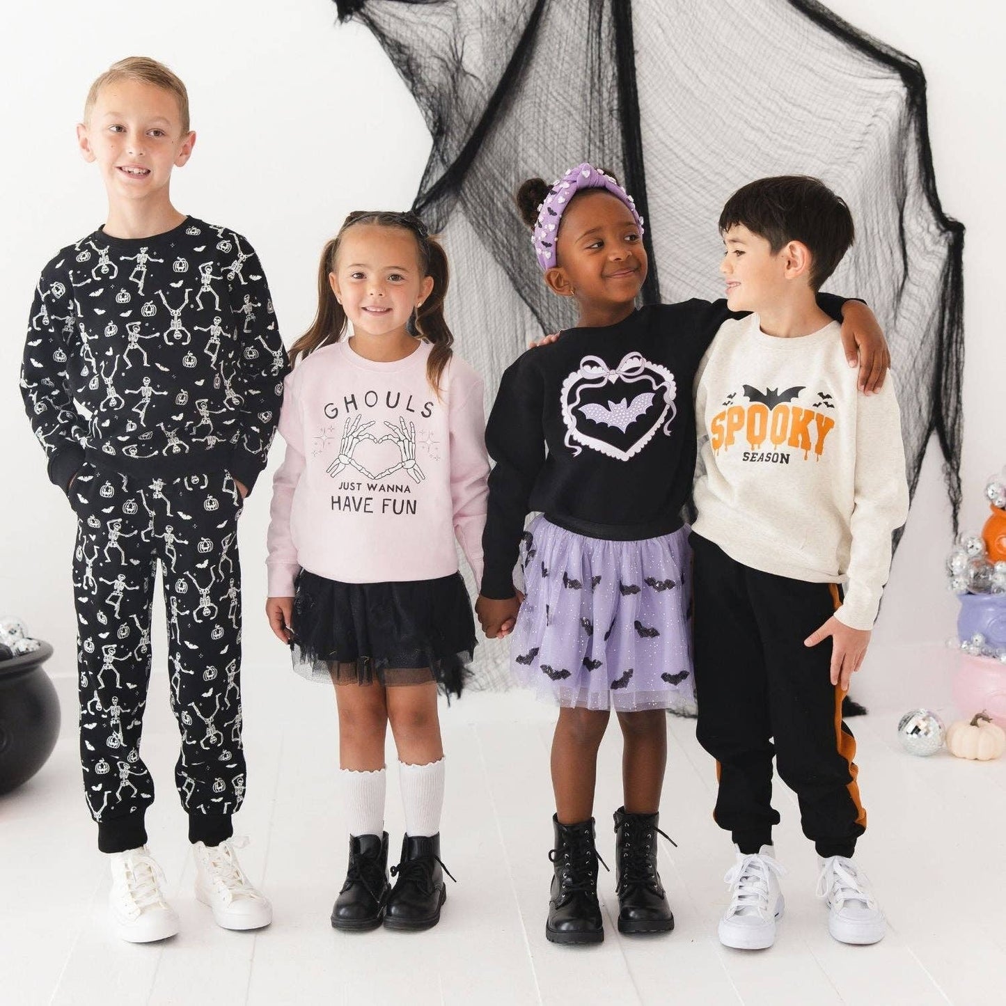 Four children standing together in a Halloween-themed studio setting, wearing matching long-sleeve sweatshirts and pants with spooky prints. The outfits feature black, white, pink, and lavender colors with bat and spider motifs, set against a white background with black netting and small decorative props.