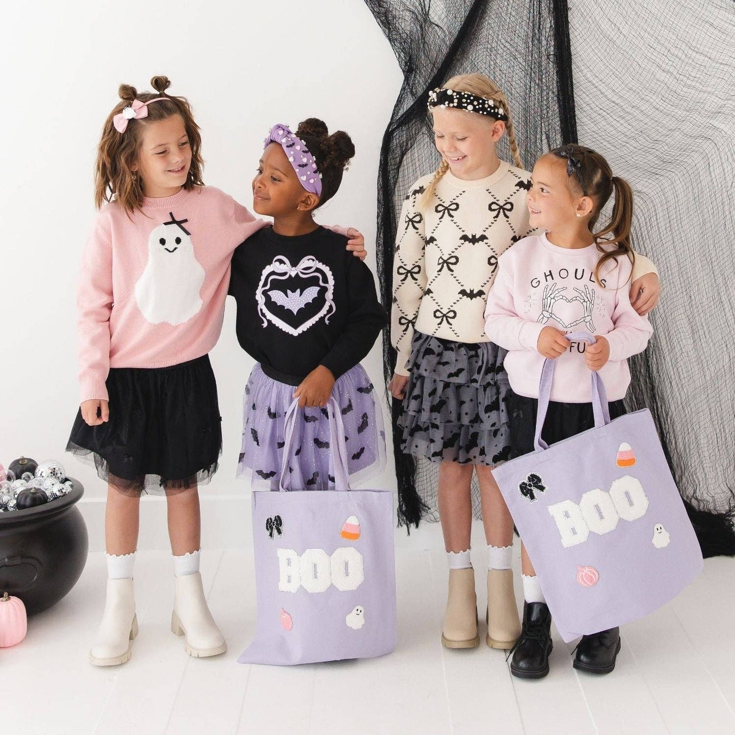 Four young girls wearing Halloween-themed sweaters and skirts stand together in a studio with black netting backdrop, each holding a lavender tote bag printed with BOO, ghosts, bats, and candy graphics.