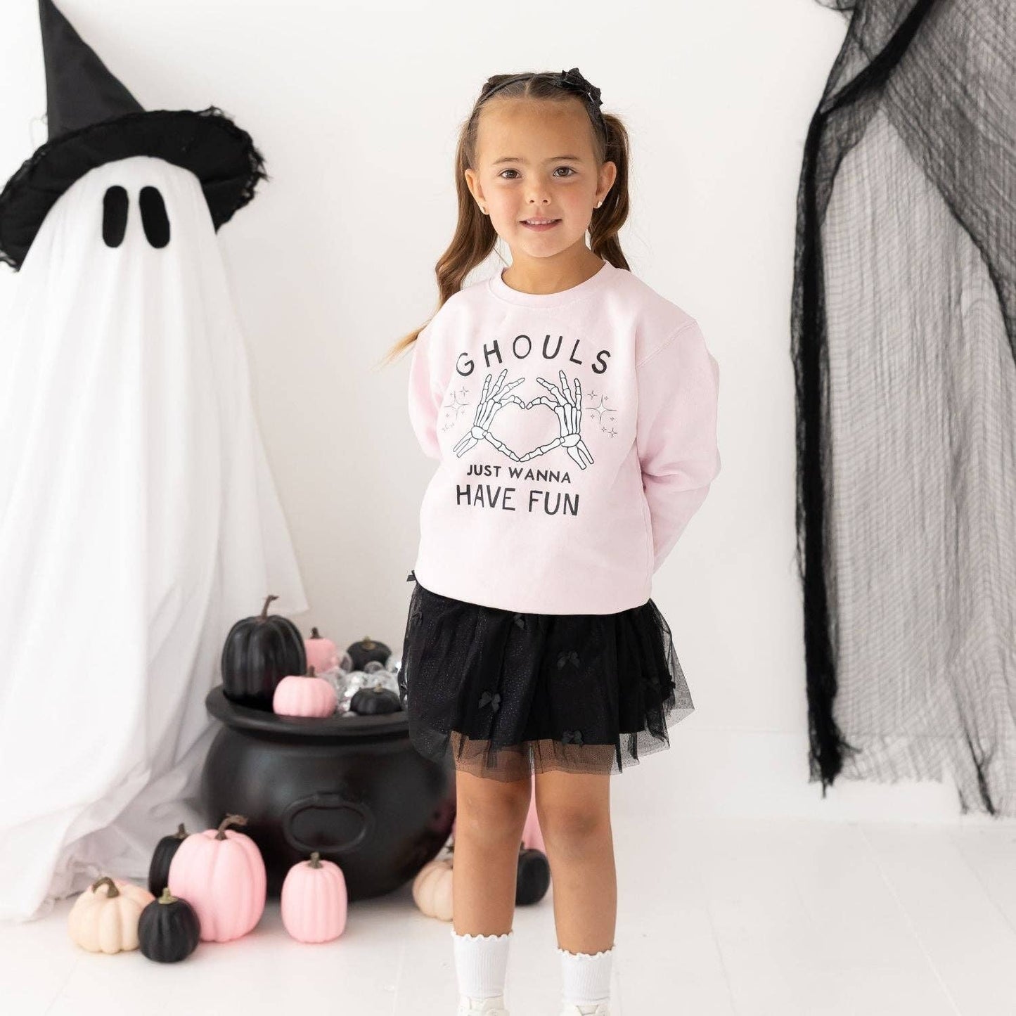 A young girl stands in a Halloween-themed studio setting wearing a light pink sweatshirt with 'Ghouls Just Wanna Have Fun' printed on the front, paired with a black tulle tutu skirt and white socks. She is posed in front of a ghost decoration, pumpkins, and black netting.