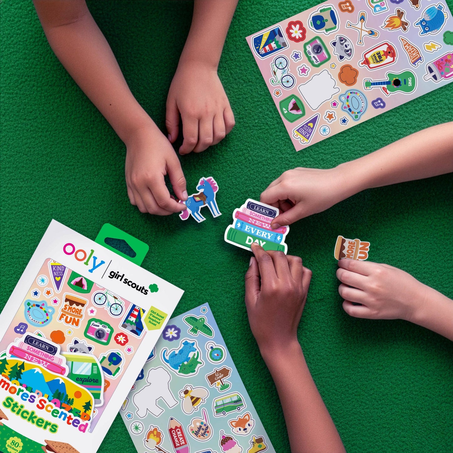Children playing with colorful stickers on a green surface, with 'ooly' and 'Girl Scouts' products visible.