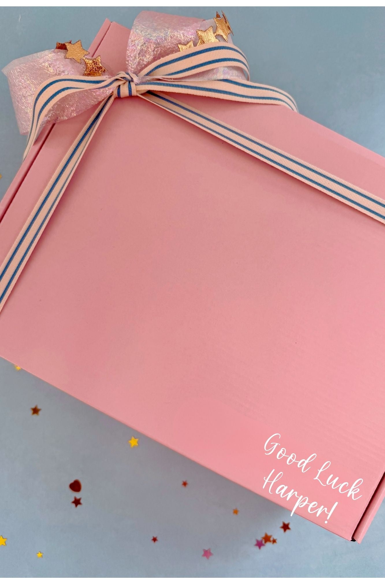A pink gift box with a matching pink ribbon bow decorated with gold star accents, placed on a light blue background with scattered confetti. White script text on the box reads 'Good Luck Harper!'.