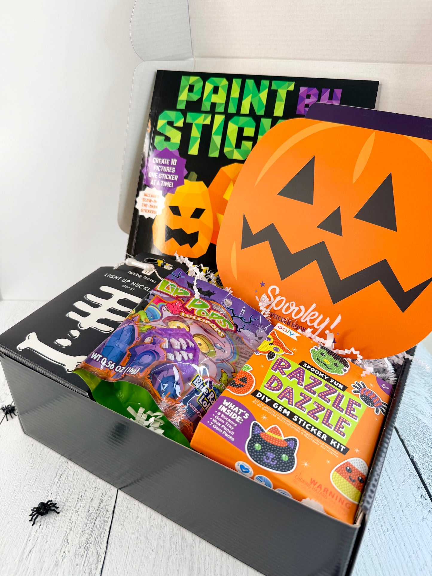 A Halloween-themed gift box filled with party supplies and snacks, including a large orange jack-o'-lantern sticker pack, a paint-by-stick kit, candy bags, and a black box with white skull graphics.