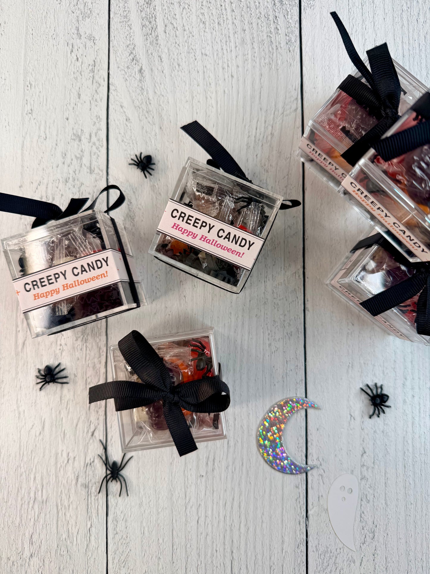 Several small clear plastic gift boxes filled with Halloween-themed candy are arranged on a white wood plank surface. Each box has a black ribbon and a label reading "Creepy Candy Happy Halloween!" with orange text.