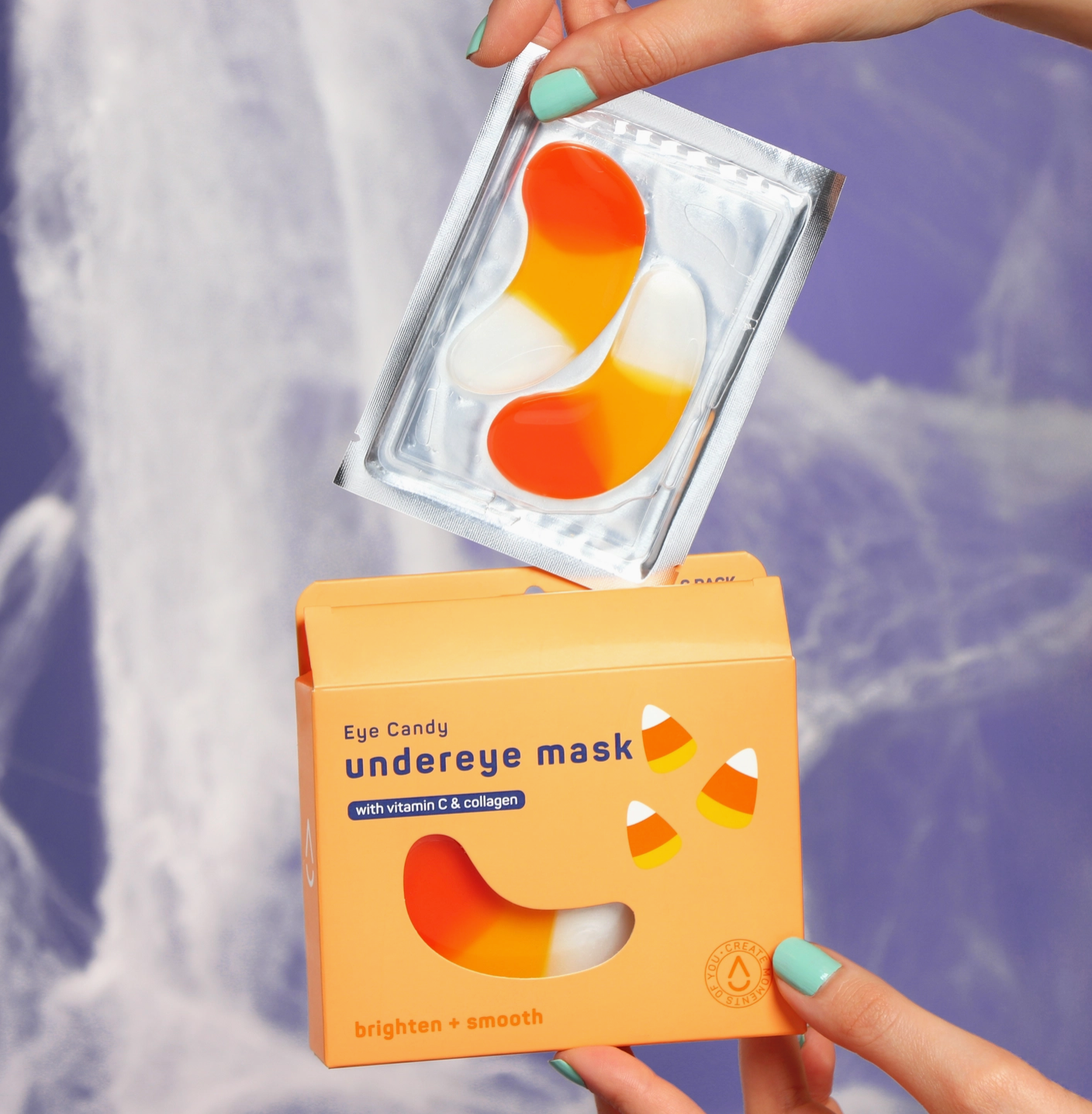 A person holds an orange box labeled 'Eye Candy underneath mask' and a clear pouch containing three eye candy shaped patch masks with orange, yellow, and white coloring, set against a blurred purple waterfall background.