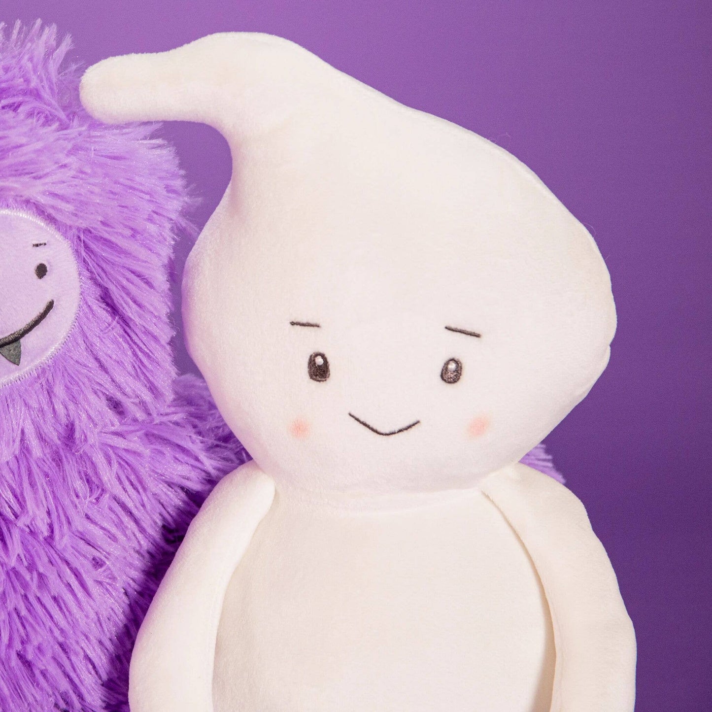 A white plush ghost toy with embroidered eyes, a smile, and pink cheeks, shown next to a purple fuzzy plush on a solid purple background.