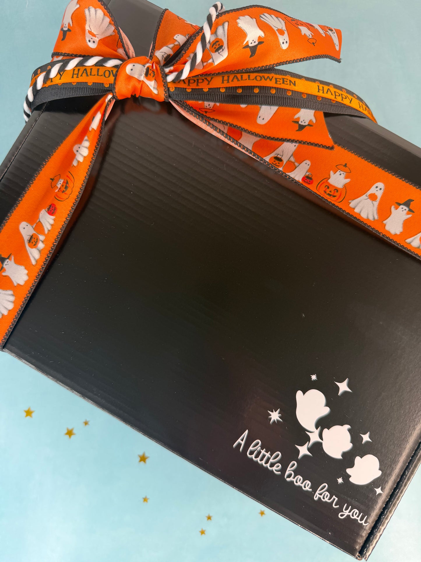 A black gift box tied with an orange and black striped ribbon printed with cartoon ghosts and the words 'Happy Halloween'. The box has white ghost illustrations and the text 'A little boo for you' on a light blue background with small gold stars.
