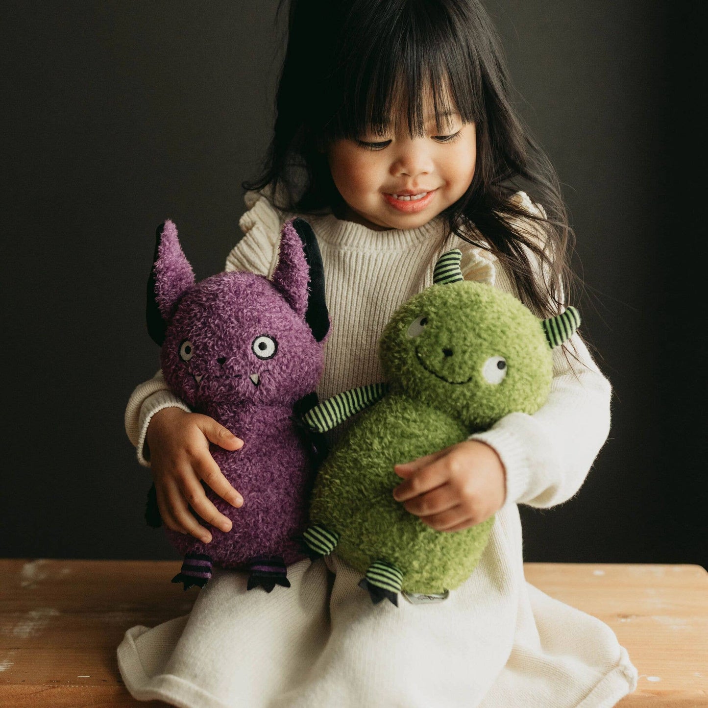 Child holding two plush toys, one purple and one green, against a dark background.