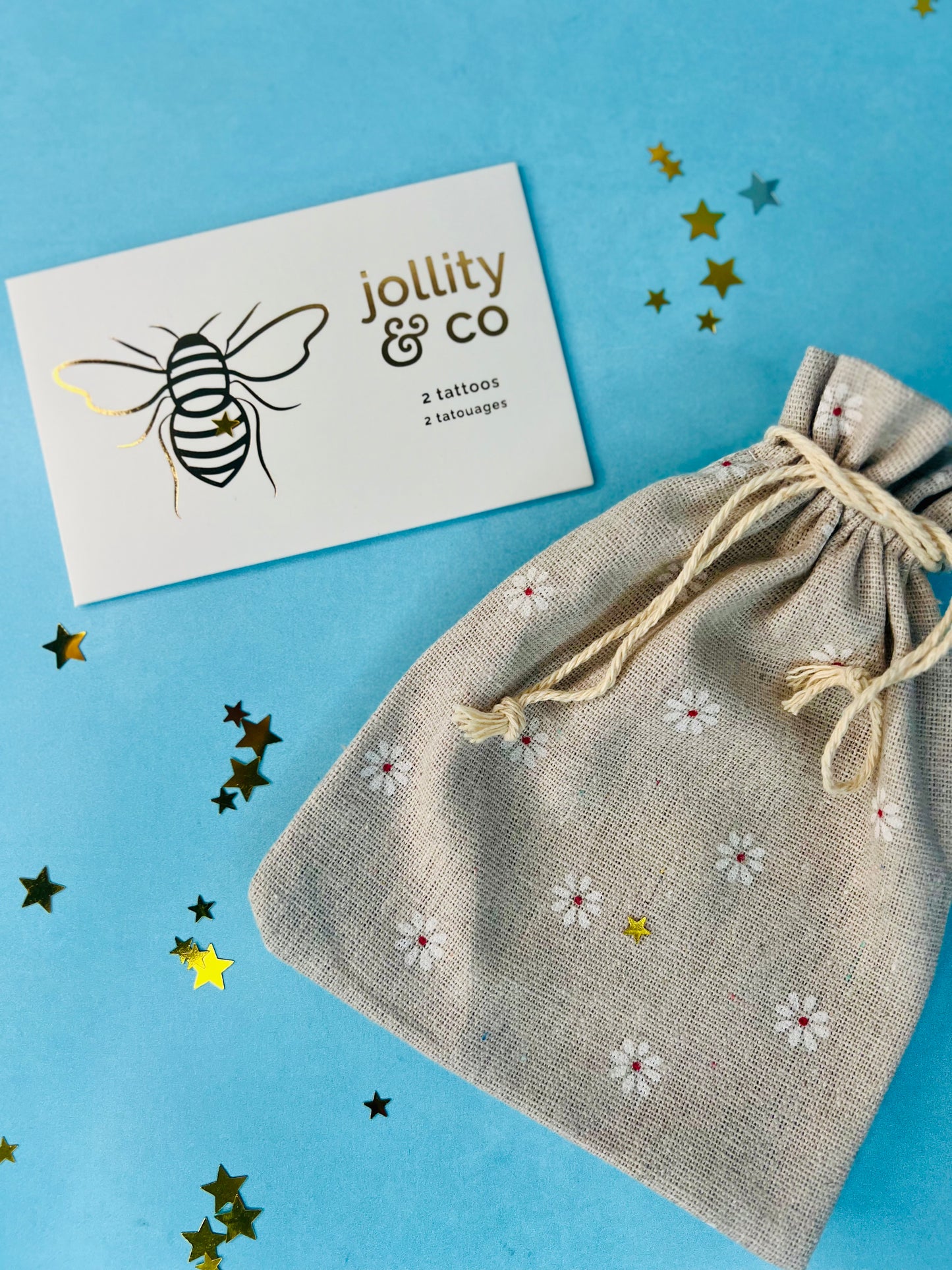 A party favor set including a burlap drawstring pouch with daisy patterns and a card with a bee illustration. Assembled party favors on a blue background with decorative stars.
