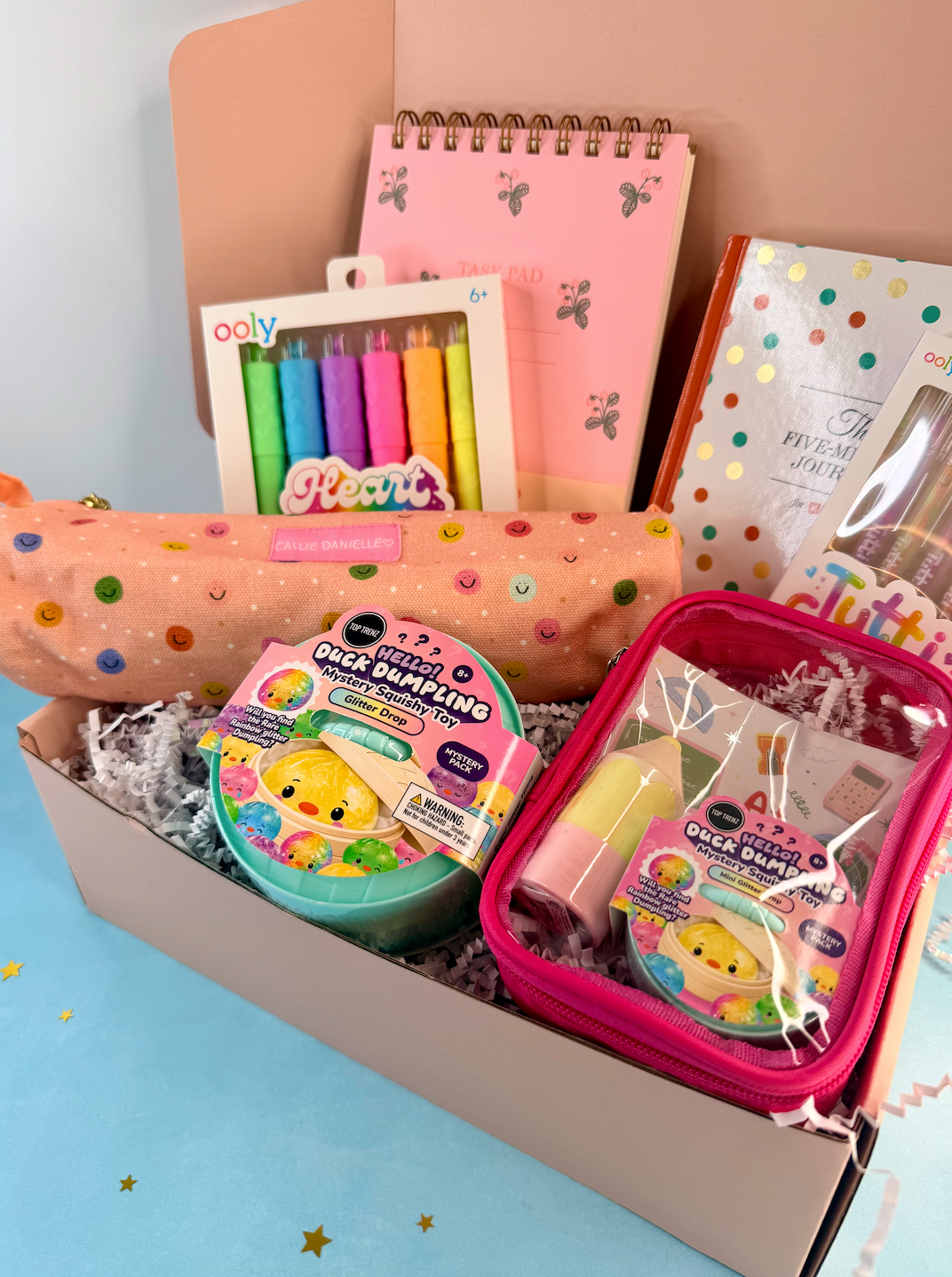 A curated gift box filled with pastel stationery and small novelty items, including a pink polka-dot pencil case, notebooks, a box of colorful gel pens, a round toy container, and a pink zip pouch with a clear front.