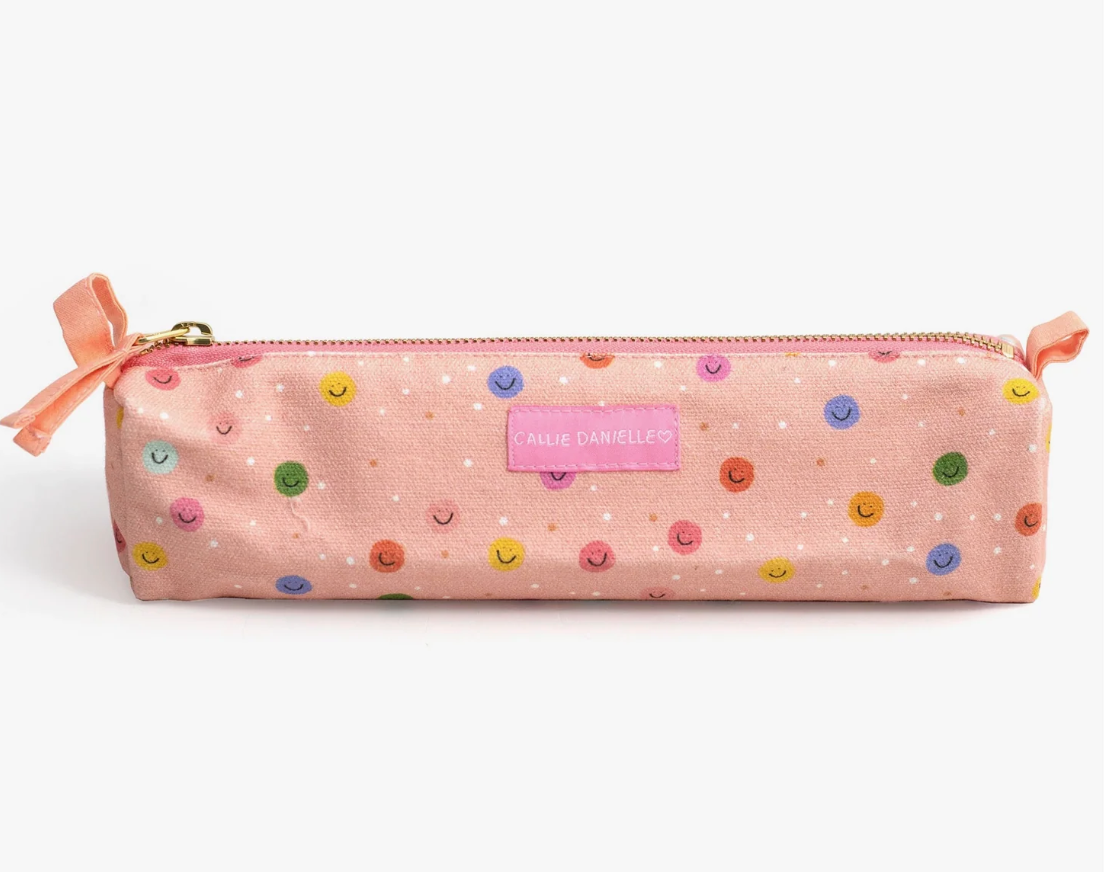 A pink fabric pencil case with a polka-dot pattern in multiple colors, featuring a small pink label that reads 'CAREY D'AMERICANS' and a peach-colored ribbon bow on the left side, set against a plain white background.