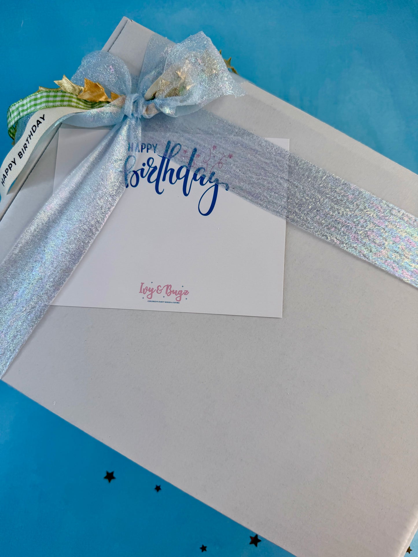 A white gift box wrapped with a large iridescent silver ribbon and a matching bow, with a small white card that says 'Happy Birthday' in blue script. The box is photographed on a bright blue background with a few small black star shapes.