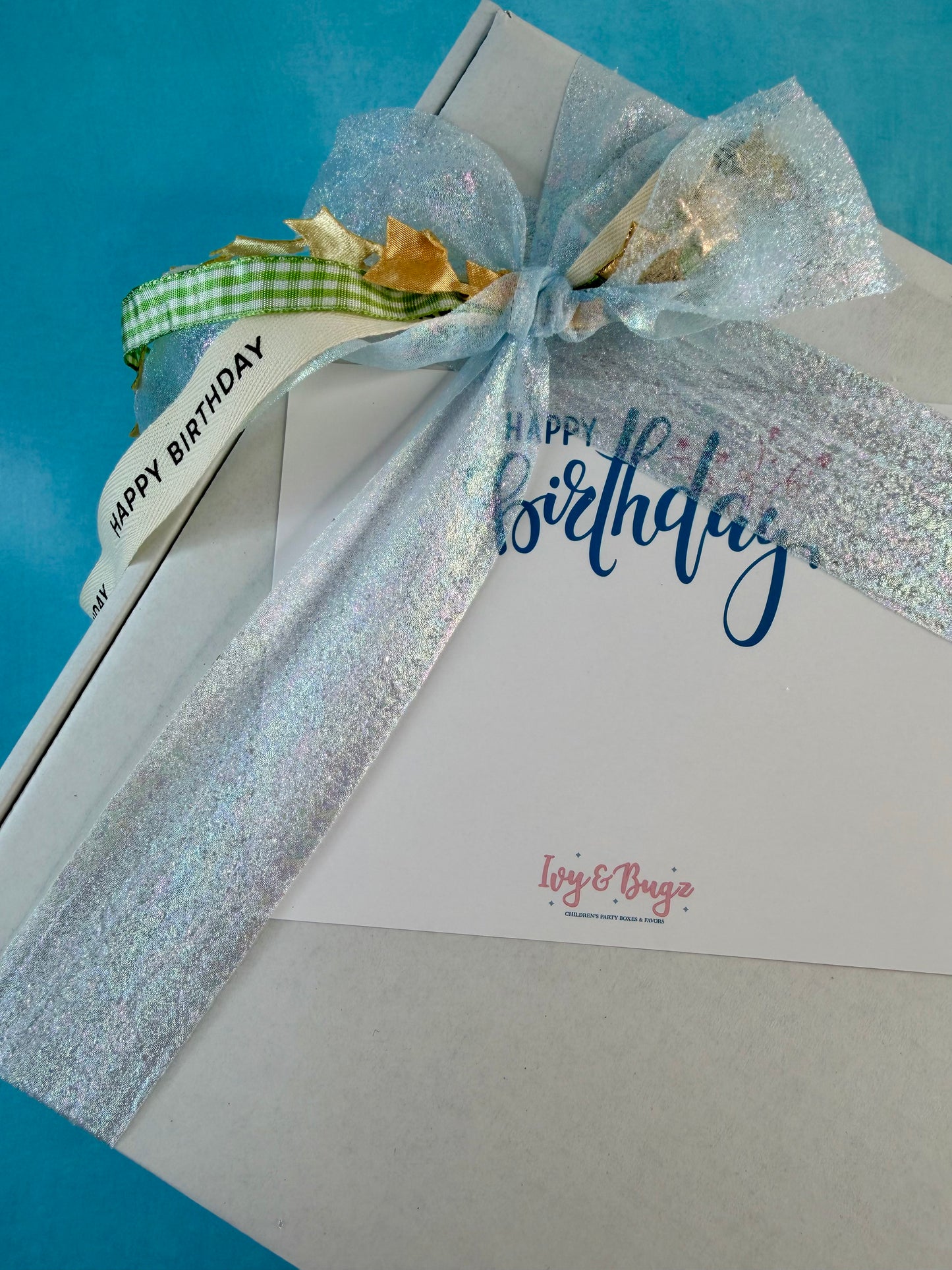 A square gift box wrapped in a large iridescent silver glitter ribbon tied in a bow, with a green gingham ribbon and gold foil accents. A white card inside reads 'Happy Birthday' in blue script and has the brand name 'Loy & Bega' printed in pink near the bottom, set against a bright blue background.